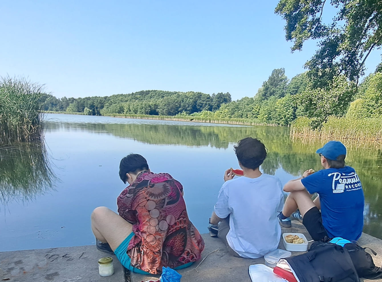 Drei Personen sitzen an einem Steg, blicken auf einen See und picknicken.