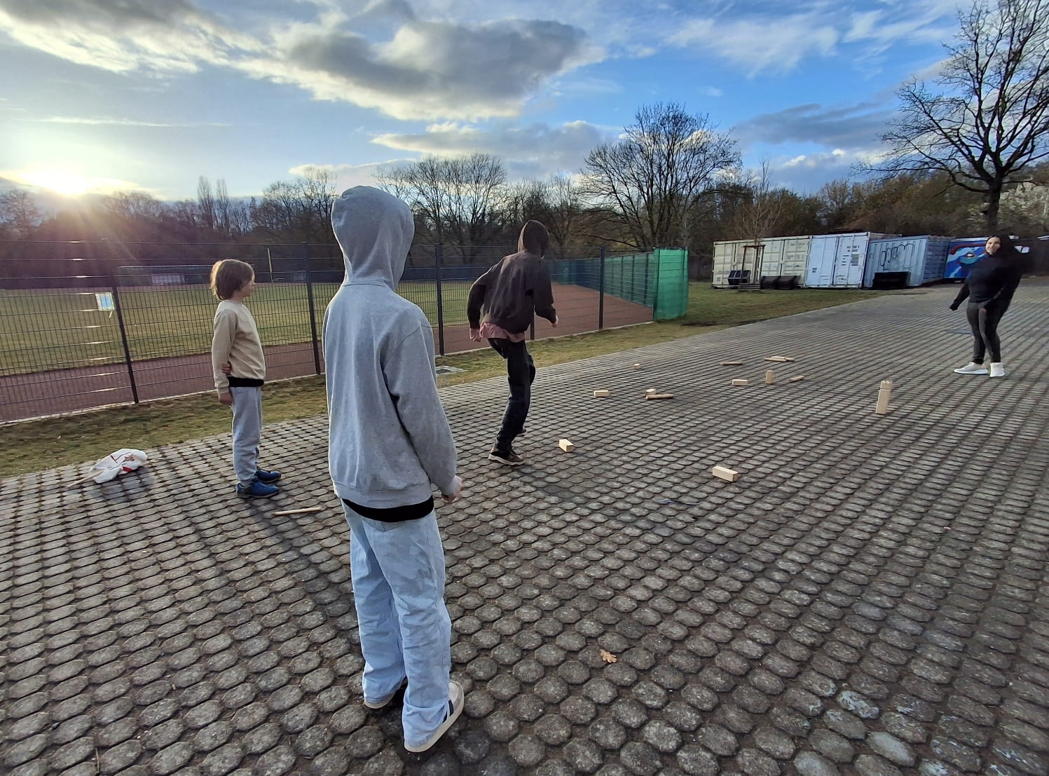 Am Rande eines Sportplatzes spielen drei Kinder Kubb.