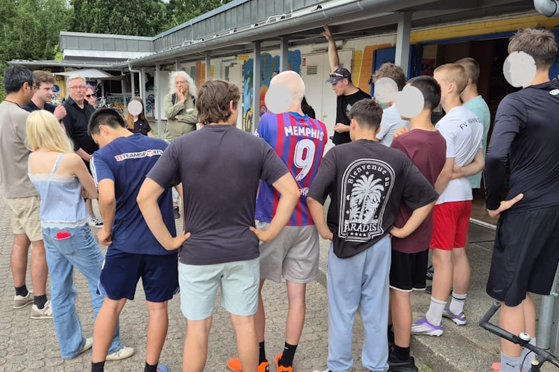 Eine Gruppe von Jugendlichen diskutiert am Rande eines Sportplatzes mit Politikern.