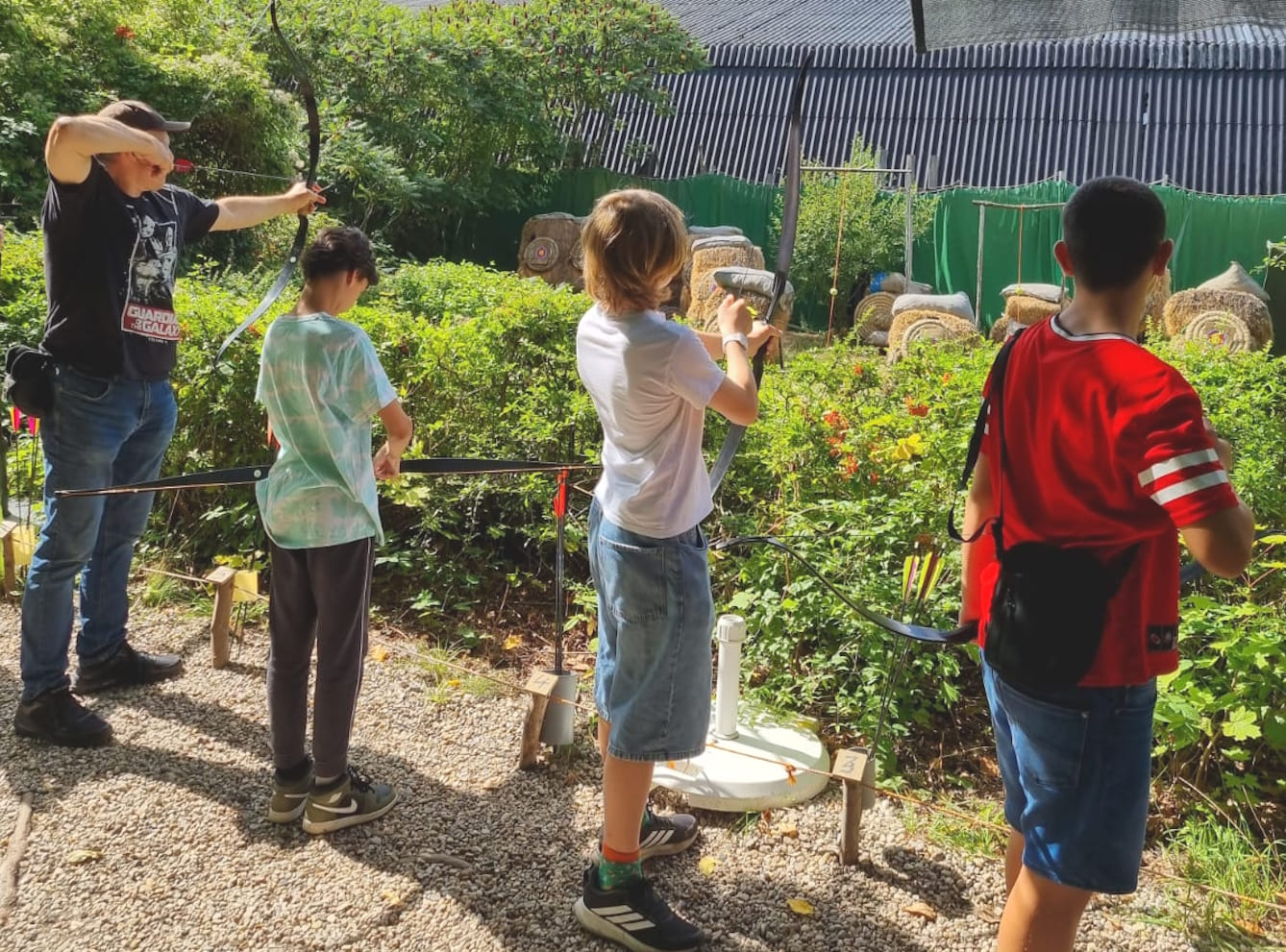 DRei Kinder und ein Betreuer stehen in einem Freigelände und Schießen mit Pfleil und Bogen.