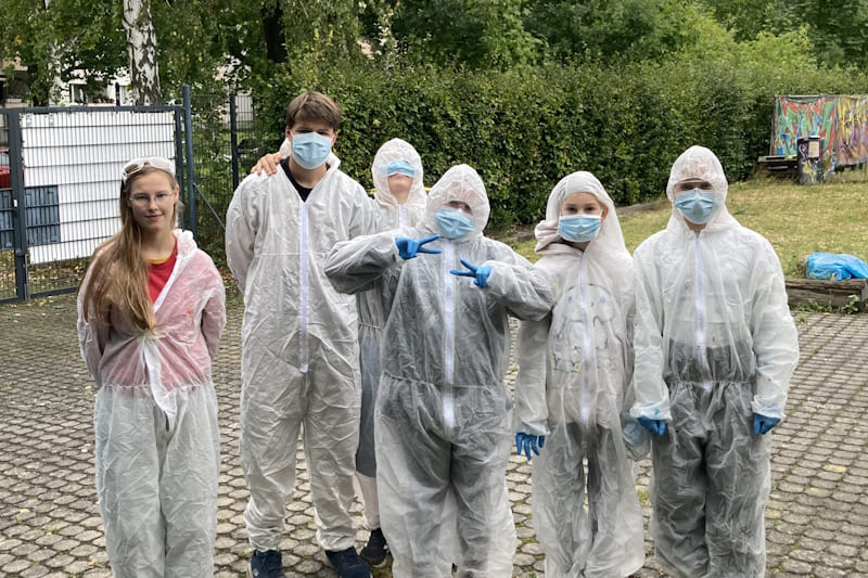Sechs junge Menschen in einer Gruppe haben weiße Schutzanzüge an und Masken aufgesetzt, um gleich Graffities zu sprühen.