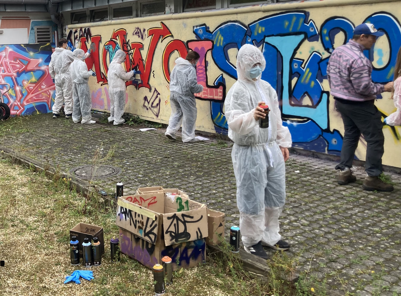 Mehrere Jugendliche in weißen Schutzanzügen sprühen Graffities an eine legale, mobile Graffiti-Wand.