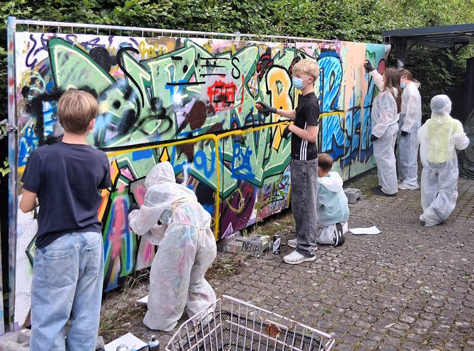 Kinder stehen in Schutzanzügen an einer Wand und sprühen Graffitis.