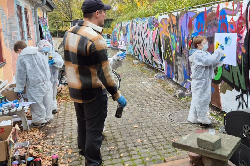 Gruppe von Jungen sprüht an eine Wand zum ersten Mal Graffities. Ein Graffiti-Künstler leitet sie an.