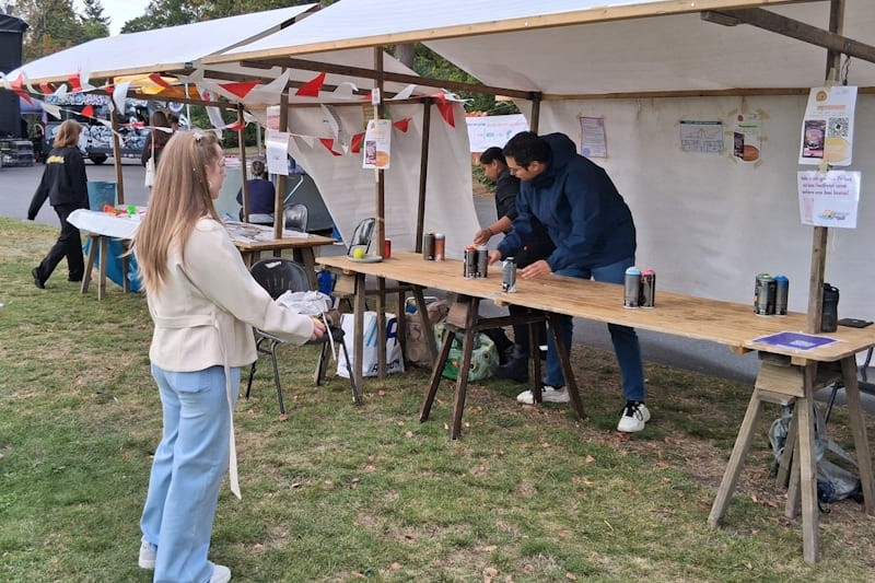 Marktstand mit aufgetürmten Spraydosen zum Abwerfen mit zwei Mitarbeitenden