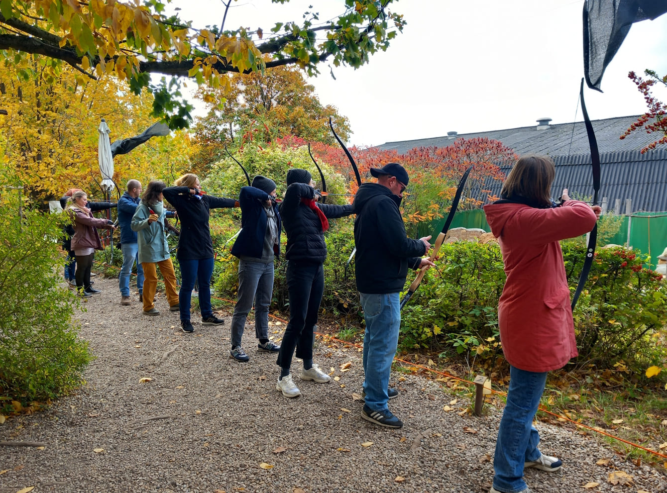 Mehrere Personen stehen an einem Schießstand mit Pfeil und Bogen umgeben von Bäumen mit buntem Herbstlaub.