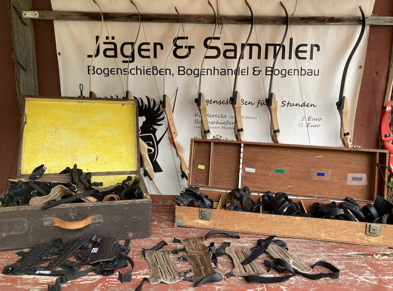 Aufgereihte Bögen, Armschützer und Handschuhe zum Bogenschießen in zwei Holzkästen auf einem Tisch.
